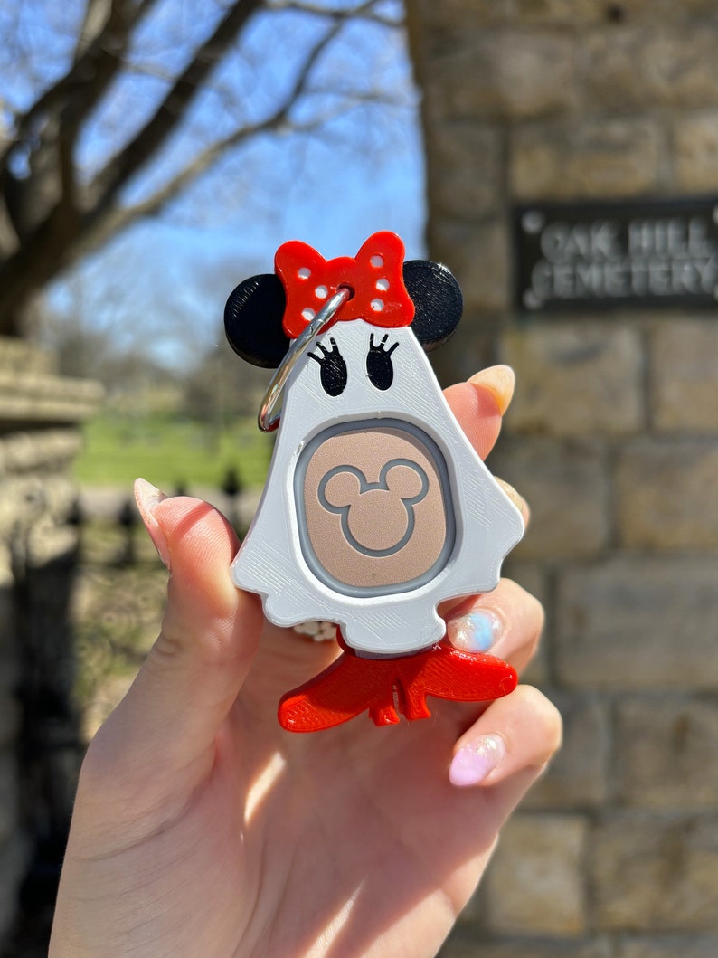 Puede incluir: Llavero blanco en forma de fantasma con un lazo rojo con lunares blancos, orejas negras y una cabeza de Mickey Mouse marr&oacute;n en la parte delantera. El llavero tiene un anillo plateado y patas rojas.