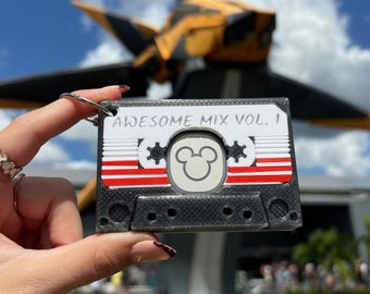 Könnte beinhalten: Ein schwarzer Kassetten-Schlüsselanhänger mit roten und weißen Streifen und einem Mickey-Mouse-Symbol. Der Text "AWESOME MIX VOL. 1" ist auf der Kassette geschrieben.