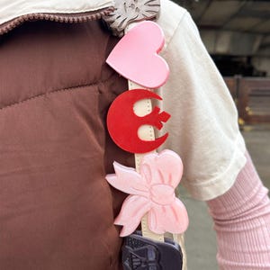 Könnte beinhalten: Ein cremefarbener Handtaschenriemen mit dekorativen Anhängern. Zu den Anhängern gehören eine graue Schleife, ein rosa Herz, ein roter Halbmond, eine rosa Schleife und ein schwarzes Darth-Vader-Design. Der Riemen ist an einer braunen Weste befestigt.
