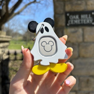 Puede incluir: Llavero blanco en forma de fantasma con orejas negras de Mickey Mouse y pies amarillos. El llavero tiene una cabeza de Mickey Mouse plateada en el centro. El llavero est&aacute; siendo sostenido por una mano.