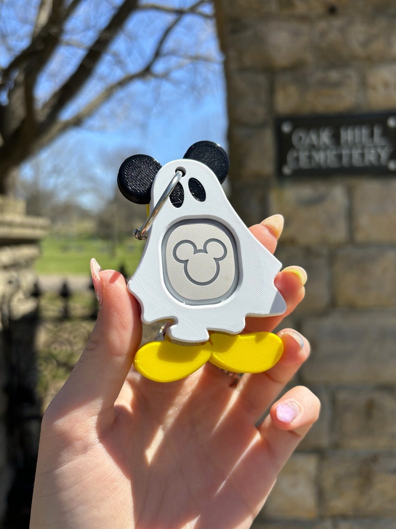 Puede incluir: Llavero blanco en forma de fantasma con orejas negras de Mickey Mouse y pies amarillos. El llavero tiene una cabeza de Mickey Mouse plateada en el centro. El llavero est&aacute; siendo sostenido por una mano.