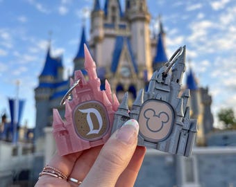 May include: Two castle-shaped keychains, one pink and one silver, featuring a Disney logo and the letter "D". The keychains are held in front of Cinderella's Castle at Walt Disney World.