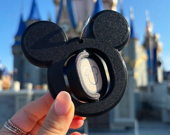 Könnte beinhalten: Schwarzer Mickey Mouse-förmiger Schlüsselanhänger mit kreisförmiger Mitte. Die Mitte hat ein helles Design mit einer Mickey Mouse-Silhouette. Der Hintergrund zeigt ein Schloss mit blauen Türmen unter klarem Himmel.