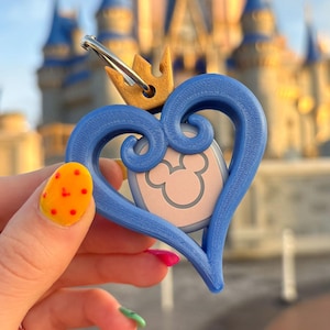 May include: A blue heart-shaped keychain with a gold crown and a silver clasp. The keychain features a pink and white Mickey Mouse design in the center. The background shows a blurred view of a castle.