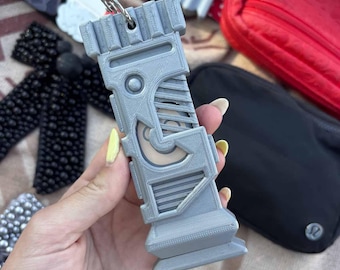 Könnte beinhalten: Ein grauer, 3D-gedruckter Schlüsselanhänger in einem geometrischen Design. Der Schlüsselanhänger hat eine rechteckige Form mit komplizierten Details und einen Metallring zum Befestigen von Schlüsseln. Weitere Accessoires sind im Hintergrund sichtbar, darunter ein roter Riemen und eine schwarze Tasche.