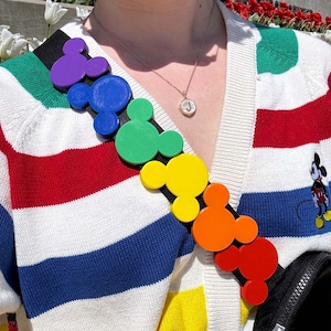May include: A rainbow-colored Mickey Mouse head strap for a bag. The strap is made of plastic and has a black adjustable strap.