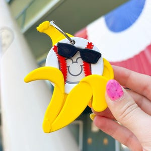Peut inclure: Un porte-clés en forme de banane jaune avec un design de baseball. La balle de baseball a des lunettes de soleil noires, des coutures rouges et une silhouette de Mickey Mouse. Le porte-clés est tenu par une personne avec du vernis à ongles rose.