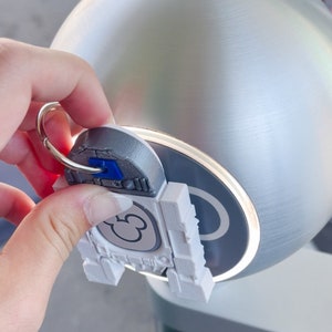 Puede incluir: Un llavero de pl&aacute;stico blanco con una cabeza de Mickey Mouse y un anillo plateado. El llavero se est&aacute; utilizando para abrir una manija de puerta plateada.