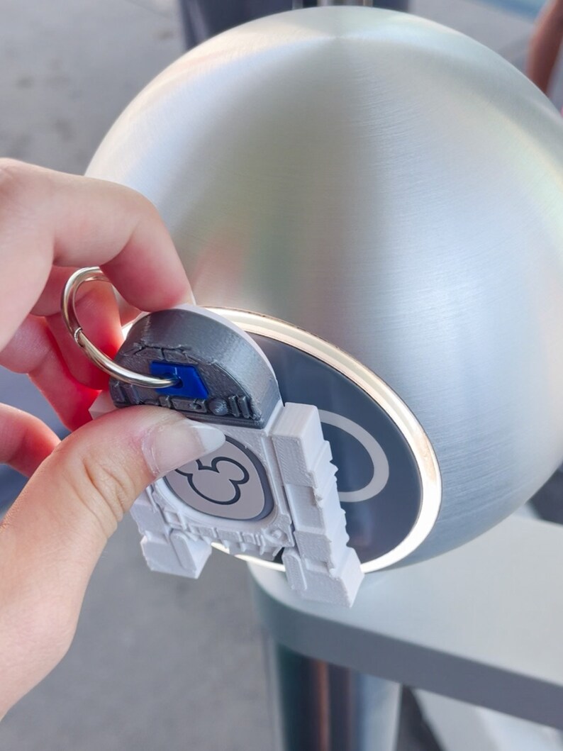 Puede incluir: Un llavero de pl&aacute;stico blanco con una cabeza de Mickey Mouse y un anillo plateado. El llavero se est&aacute; utilizando para abrir una manija de puerta plateada.