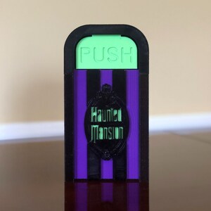May include: A black and purple Haunted Mansion themed doorstop with a green "PUSH" button.