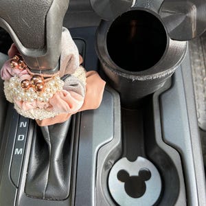 May include: Black car cup holder with a Mickey Mouse design. The cup holder has a black lid with Mickey Mouse ears and a white Mickey Mouse head on the bottom.