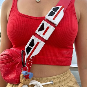 Peut inclure: Un débardeur rouge avec une bandoulière de monorail blanche et rouge et un sac à bandoulière rouge avec un porte-clés en forme d'ours brun.