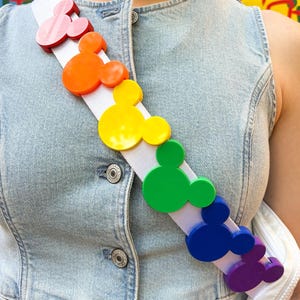 Könnte beinhalten: Ein weißer Umhängeriemen mit bunten Mickey Mouse-Kopf-Formen in einem Regenbogenmuster. Der Riemen wird über einer Jeansweste getragen. Die Mickey Mouse-Formen sind rot, orange, gelb, grün, blau und lila.