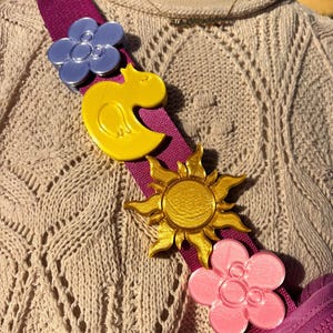 May include: A purple crossbody bag strap featuring 3D-printed charms in various colours. Charms include a blue flower, a yellow duck, a gold sun, and a pink flower. The strap is attached to a matching purple bag.