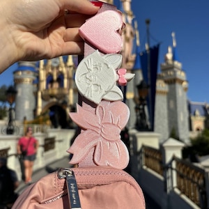 May include: A pink crossbody bag with a zipper closure and a silver zipper pull. The bag has three charms attached to the strap: a pink heart, a white cat head, and a pink bow.