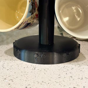 May include: A black mug tree stand with a circular base featuring embossed mouse ear silhouettes. The stand has a central vertical post and holds multiple ceramic mugs, including one with a colorful graphic print and a plain white interior.