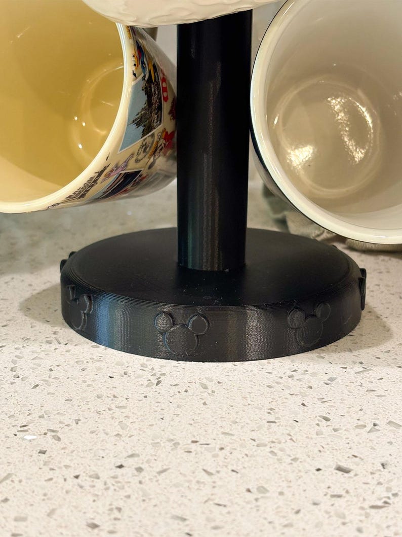 May include: A black mug tree stand with a circular base featuring embossed mouse ear silhouettes. The stand has a central vertical post and holds multiple ceramic mugs, including one with a colorful graphic print and a plain white interior.