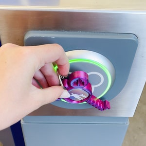 Puede incluir: Un llavero de color morado con forma de Mickey Mouse con un anillo plateado se utiliza para abrir una cerradura de puerta electr&oacute;nica gris y plateada. La cerradura tiene una luz verde alrededor del borde.