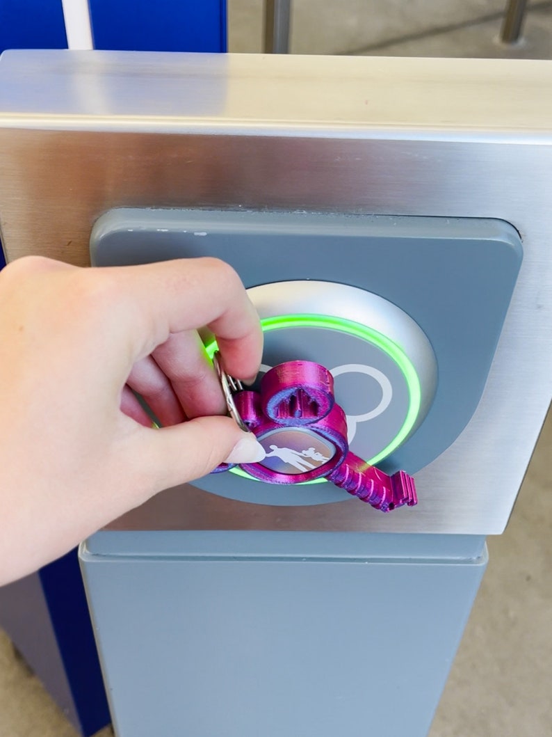 Puede incluir: Un llavero de color morado con forma de Mickey Mouse con un anillo plateado se utiliza para abrir una cerradura de puerta electr&oacute;nica gris y plateada. La cerradura tiene una luz verde alrededor del borde.