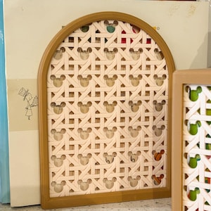 May include: A decorative arched panel with a woven design featuring Mickey Mouse silhouettes. The panel has a light brown wooden frame and a cream-colored woven pattern. The panel is placed in front of a stack of books.