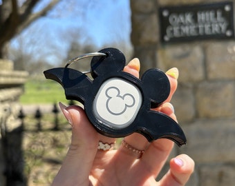 Puede incluir: Llavero de plástico negro en forma de murciélago con un logotipo blanco de cabeza de Mickey Mouse y un anillo plateado. El llavero está siendo sostenido por una mano frente a una pared de piedra con un letrero que dice "Oak Hill Cemetery."