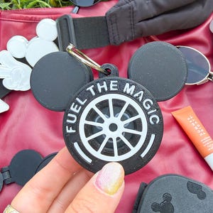 May include: A black circular keychain featuring two rounded ears at the top and a white steering wheel graphic in the center. The text FUEL THE MAGIC is printed in white capital letters along the upper edge of the circular frame, which includes a metal ring.