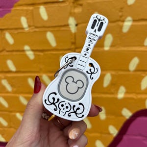 May include: White guitar-shaped keychain with a Mickey Mouse head on the front and a skull design on the back. The keychain is decorated with black and white details.
