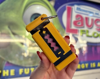 Puede incluir: Llavero de plástico amarillo y negro con forma de caja pequeña con un diseño rosa y blanco en el interior. El llavero tiene un anillo plateado.