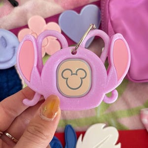 May include: A light purple keychain featuring a cartoon character design with large ears and a Mickey Mouse emblem. The keychain has a silver ring and is surrounded by other accessories in shades of pink and purple.