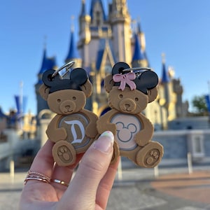 May include: Two brown teddy bear keychains with Mickey Mouse ears. One keychain has a pink bow and the other has a black and white 'D' on the front. The keychains are held in front of Cinderella's Castle at Walt Disney World.
