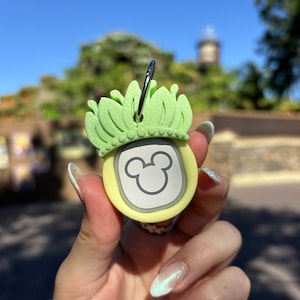 May include: A yellow and green Mickey Mouse keychain with a crown-shaped top. The keychain has a silver Mickey Mouse head on the front.