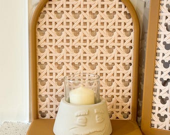May include: A tan-colored shelf with a decorative backing featuring a Mickey Mouse pattern. A light gray candle holder with a face design holds a lit candle in a glass. A matching frame is on the right.