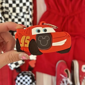 May include: Red plastic keychain shaped like the character Lightning McQueen from the Disney Pixar movie Cars. The keychain features a Mickey Mouse head in the centre and the number 95 on the side.