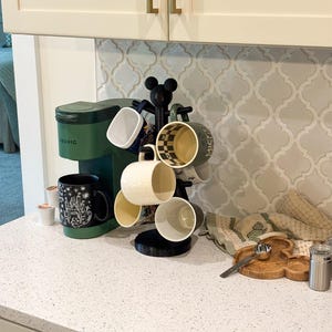 May include: A matte black mug tree with a Mickey Mouse silhouette topper holding six assorted ceramic mugs, positioned next to a forest green Keurig coffee maker. The mugs feature various patterns including checkered, solid, and graphic designs.