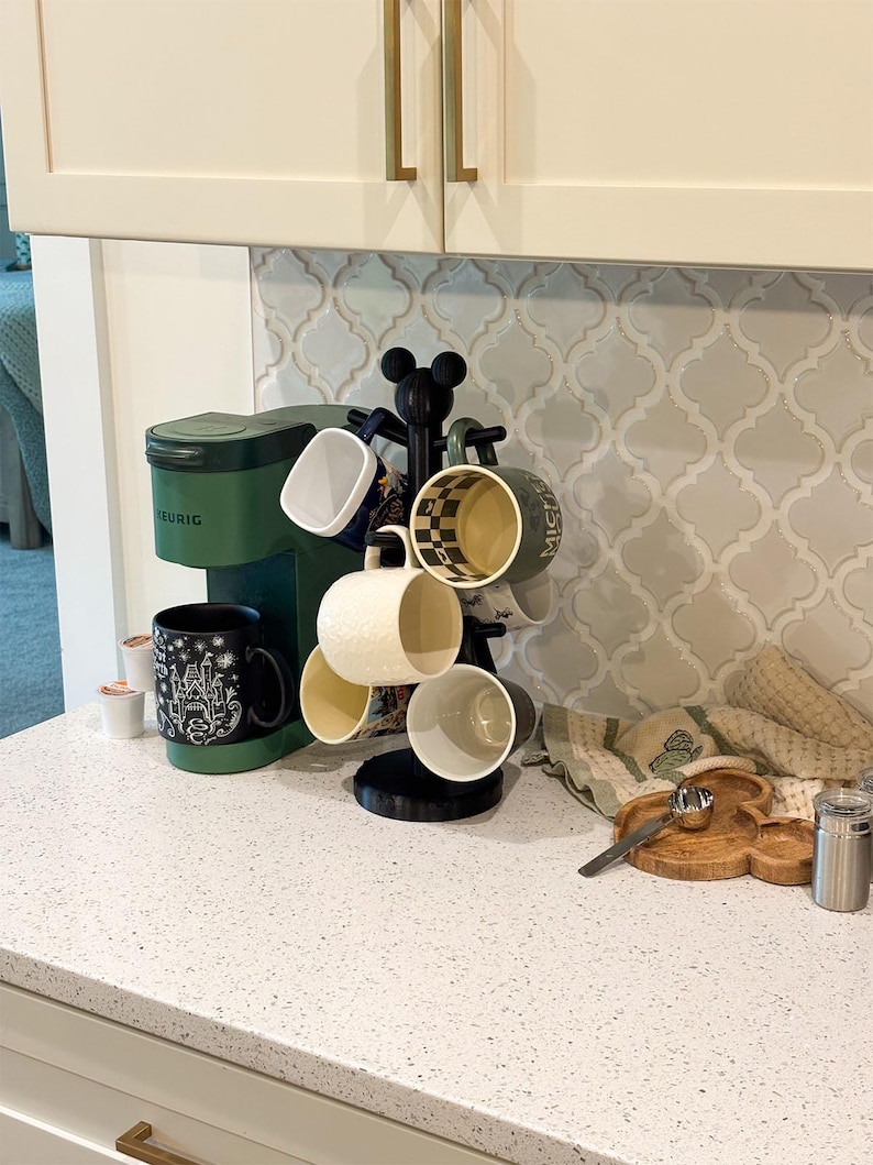 May include: A matte black mug tree with a Mickey Mouse silhouette topper holding six assorted ceramic mugs, positioned next to a forest green Keurig coffee maker. The mugs feature various patterns including checkered, solid, and graphic designs.