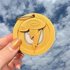 May include: A gold-colored keychain with a lightning bolt design. The keychain features a circular shape with a cloud and lightning bolt graphic. The background is a cloudy sky.