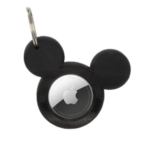 May include: Black AirTag holder shaped like Mickey Mouse ears with a silver Apple logo and text that reads "Designed by Apple in California. Assembled in China. Ultra Wideband. AirTag."