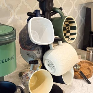 May include: A black mug tree stand featuring a Mickey Mouse ear silhouette at the top and a circular base. The stand holds several ceramic mugs, including a dark blue mug, a green mug with a checkered interior, and a white mug with embossed icons.