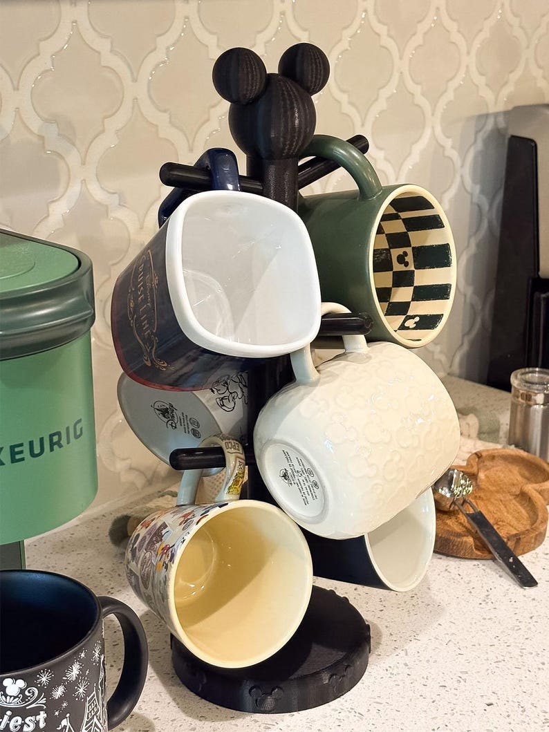 May include: A black mug tree stand featuring a Mickey Mouse ear silhouette at the top and a circular base. The stand holds several ceramic mugs, including a dark blue mug, a green mug with a checkered interior, and a white mug with embossed icons.