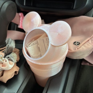 May include: A pink car trash can with Mickey Mouse ears. The trash can is made of a soft, velvety material and has a white liner. The trash can is sitting in the cup holder of a car.