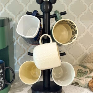May include: A black mug tree stand with a circular base and a top finial shaped like a mouse head silhouette. The stand features multiple horizontal arms designed to hold mugs, with one white textured mug currently hanging on the center arm.