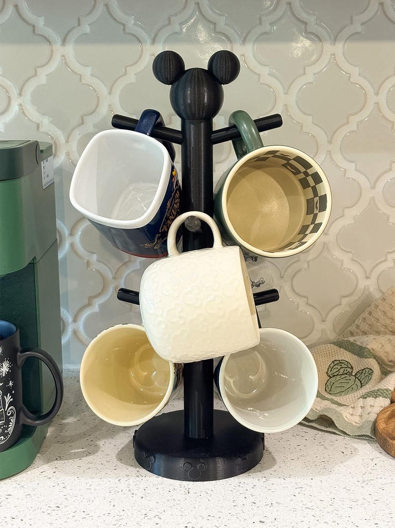 May include: A black mug tree stand with a circular base and a top finial shaped like a mouse head silhouette. The stand features multiple horizontal arms designed to hold mugs, with one white textured mug currently hanging on the center arm.