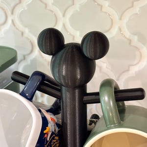 May include: A black mug tree featuring a central post topped with a three-dimensional mouse head silhouette. The stand has horizontal arms extending from the center to hold multiple mugs, including a blue mug with Disney Fantasia text.