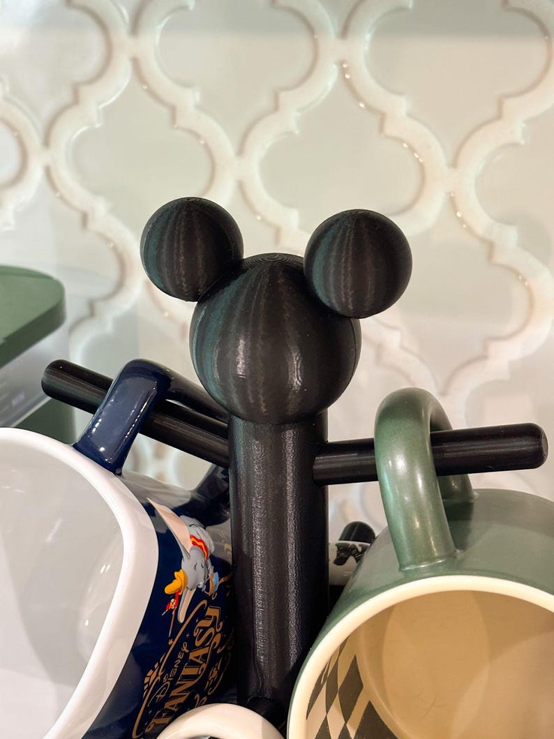 May include: A black mug tree featuring a central post topped with a three-dimensional mouse head silhouette. The stand has horizontal arms extending from the center to hold multiple mugs, including a blue mug with Disney Fantasia text.