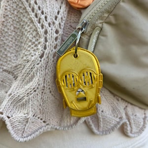 May include: A gold-colored C-3PO head keychain is attached to a beige Lululemon bag. The keychain is made of plastic and has a key ring. Other small, colorful Star Wars-themed items are visible in the background.