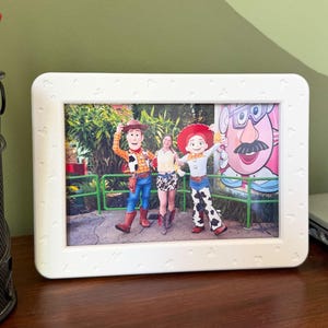 May include: A rectangular white picture frame with a subtle embossed pattern of small Mickey Mouse heads along the border. The frame holds a photograph featuring two costumed characters, Woody and Jessie, standing beside a person in a park setting.
