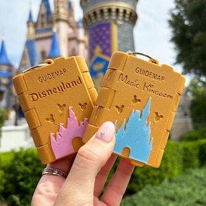 以下が含まれることがあります： 「Disneyland」とピンクの城、「Magic Kingdom」と青い城が描かれた2つの黄色のガイドブックキーホルダー。どちらもシルバーのキーリングが付いており、背景にはぼやけたお城が写っています。