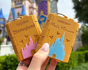 Könnte beinhalten: Zwei gelbe Schlüsselanhänger in Form von Reiseführern, einer mit der Aufschrift "Disneyland" und einem rosa Schloss, der andere "Magic Kingdom" mit einem blauen Schloss. Beide haben einen silbernen Schlüsselring und werden vor einem verschwommenen Schloss-Hintergrund gehalten.