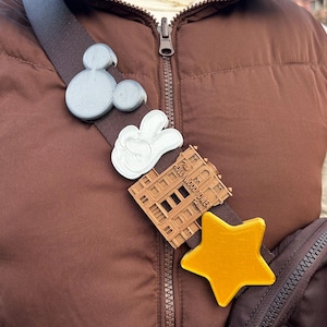 May include: A brown shoulder strap featuring decorative pins. The pins include a grey Mickey Mouse head, a white peace sign, a brown building, and a yellow star. The strap is attached to a brown vest.