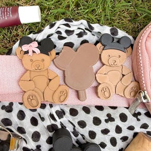 May include: Two tan teddy bear figures with black mouse-ear headbands, one featuring a small pink bow, and a central brown mouse-shaped popsicle figure. These items are positioned on a white fabric with black spots and a light pink strap.